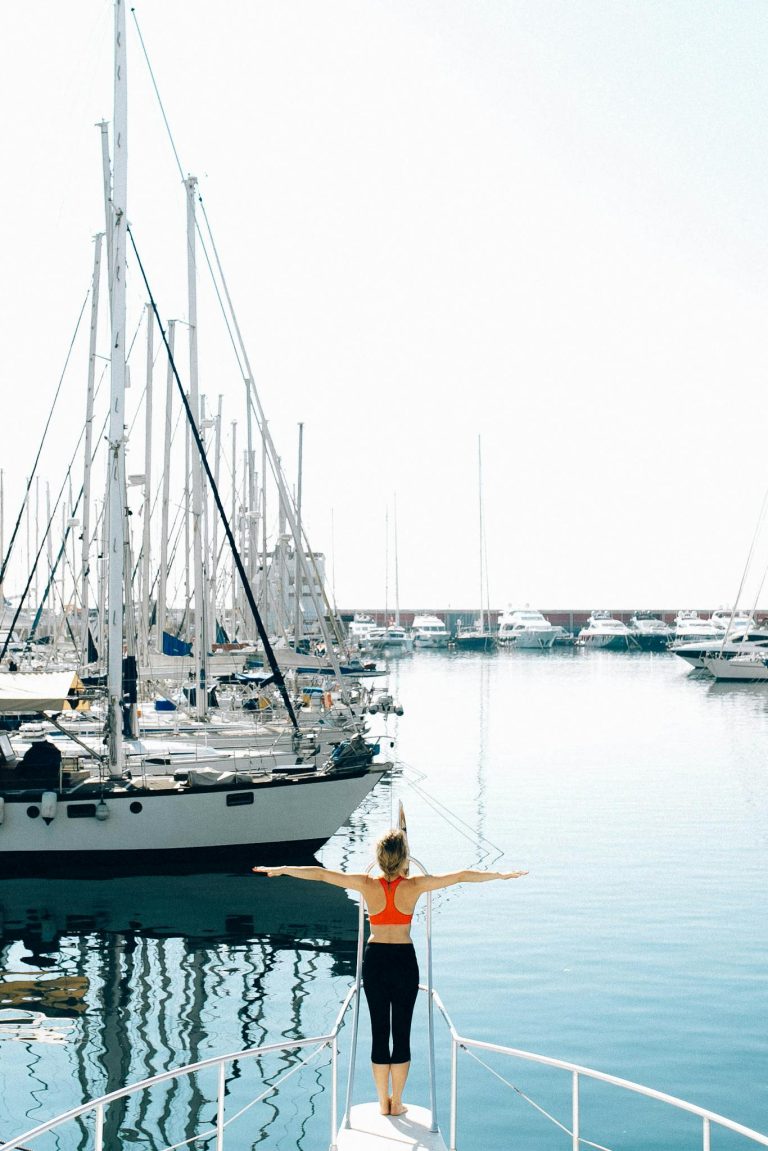 Young woman stretching on a yacht in a sunny harbor surrounded by boats and calm sea.
